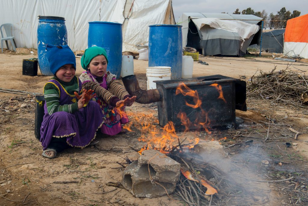 Two children are sitting near a firepit in a refugee camp