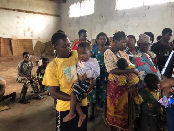 A boy is carrying a baby in a room where people are standing