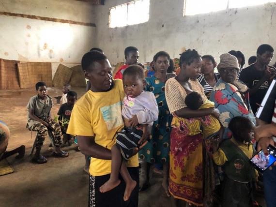 A boy is carrying a baby in a room where people are standing 