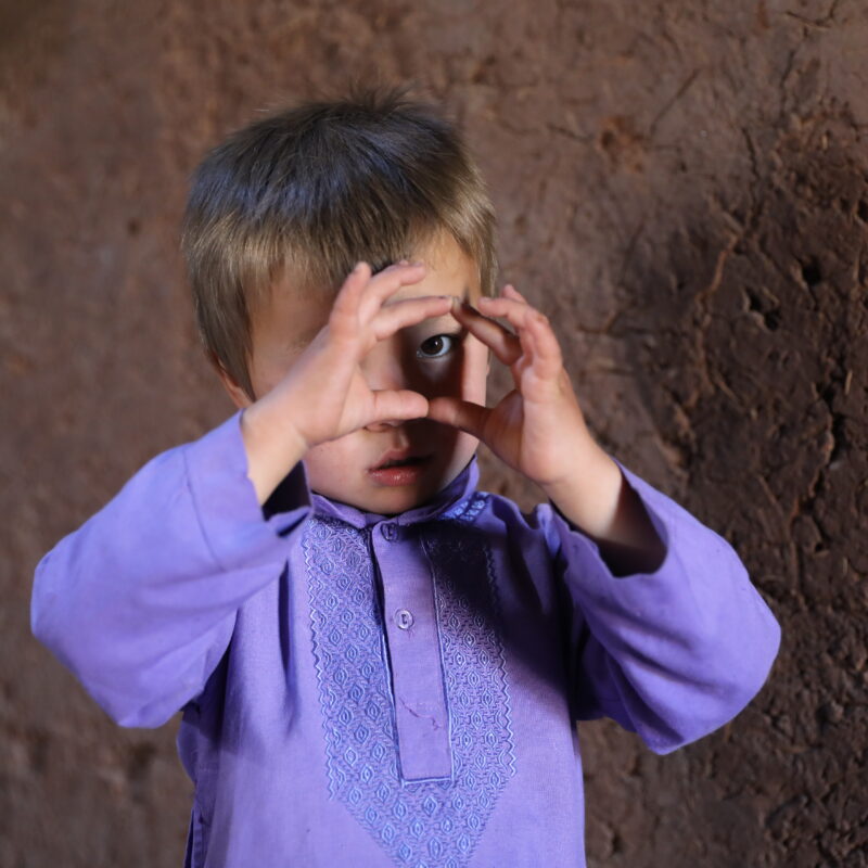 Bin Yamin (3) makes a heart with his hands as he plays, while his mother Nikbakht prepares a meal using vegetables grown locally around her home in Afghanistan.